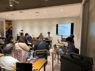 会場で岡田先生とならび、座りながらマイクを持つ西岡教授