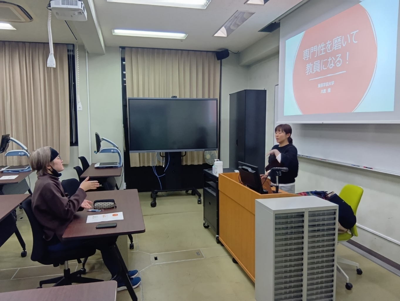 大鹿先生が、教室でパワーポイントのスクリーンを背に、学生と手話を使って話をしているところ