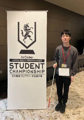 「STUDENT CHAMPIONSHIP」と書かれた高さ2ｍほどのタペストリーの向かって右横に白杖を携えて立つ酒寄寛也さんの写真
